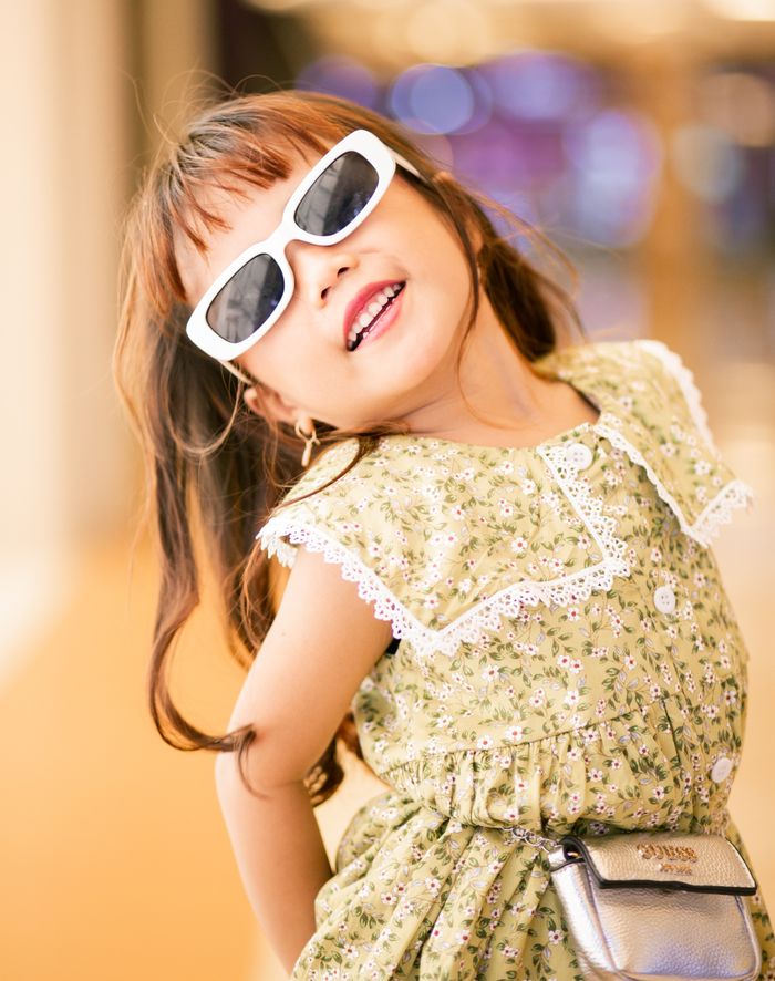 Adorable child posing indoors wearing sunglasses and a floral dress.