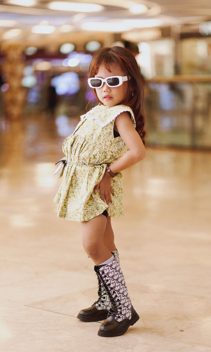 A young girl posing confidently in a shopping mall wearing a fashionable yellow dress and sunglasses.