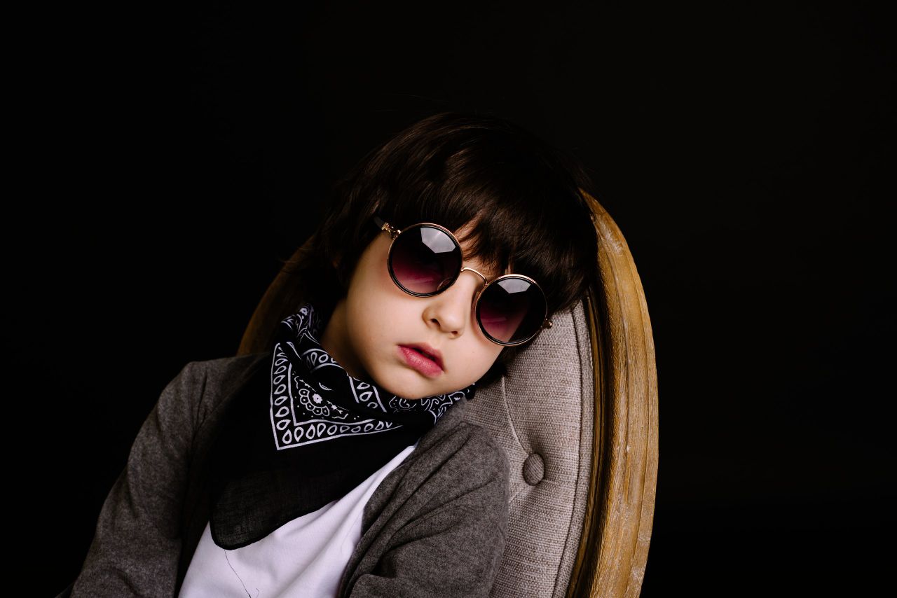 Portrait of a fashionable child wearing sunglasses and a scarf, sitting on a chair against a dark background.